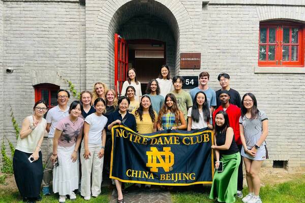 The Notre Dame Club of China-Beijing poses with their banner in front of a gray brick building with red doors and windows.