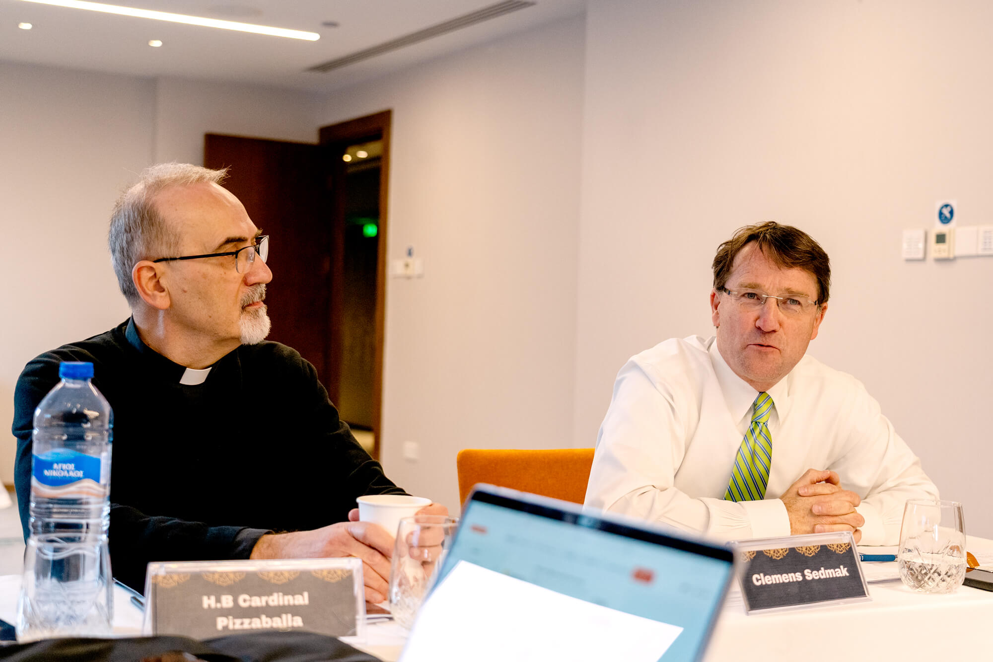 H.B. Cardinal Pizzaballa, in a black clerical shirt, looks left at a table. Next to him, Clemens Sedmak, in a white shirt and green-striped tie, clasps his hands and looks forward.