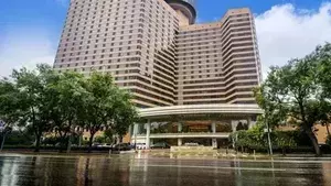 Four Seasons Hotel Beijing. Tall, tan building with numerous horizontal windows and a large, curved canopy entrance. A wet street reflects the building and surrounding green trees under a bright, cloudy sky.