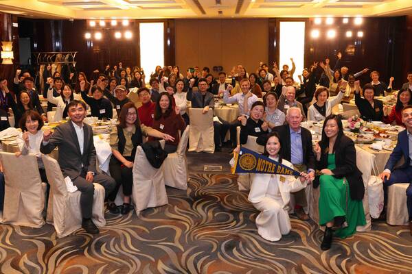 Enthusiastic group raises fists at a banquet. Woman kneels, holding a blue and gold Notre Dame pennant in front.