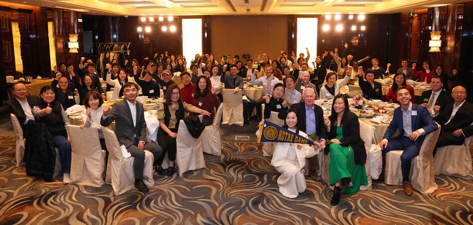 Enthusiastic group raises fists at a banquet. Woman kneels, holding a blue and gold Notre Dame pennant in front.