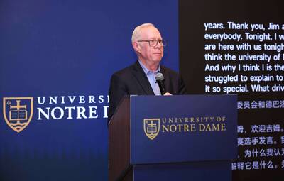 A man in a dark suit and glasses speaks at a blue University of Notre Dame podium with logos, against a blue wall and text screen.